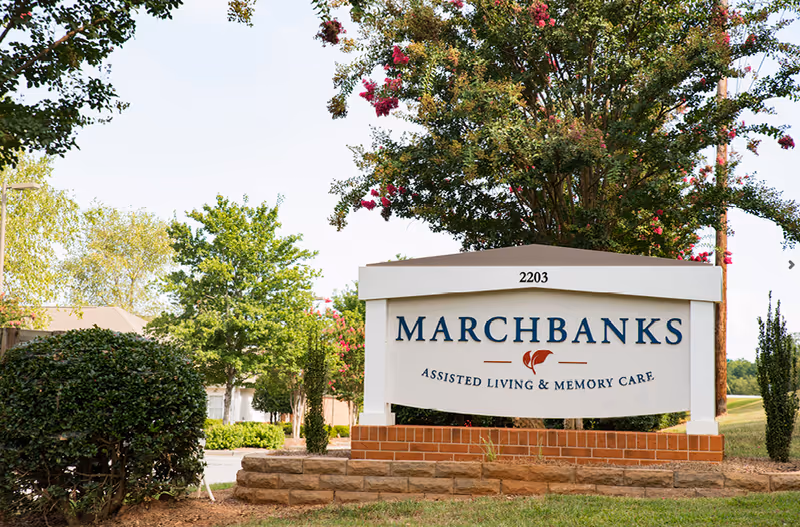 Entrance sign that reads "Marchbanks Assisted Living & Memory Care" surrounded by landscaping and trees.