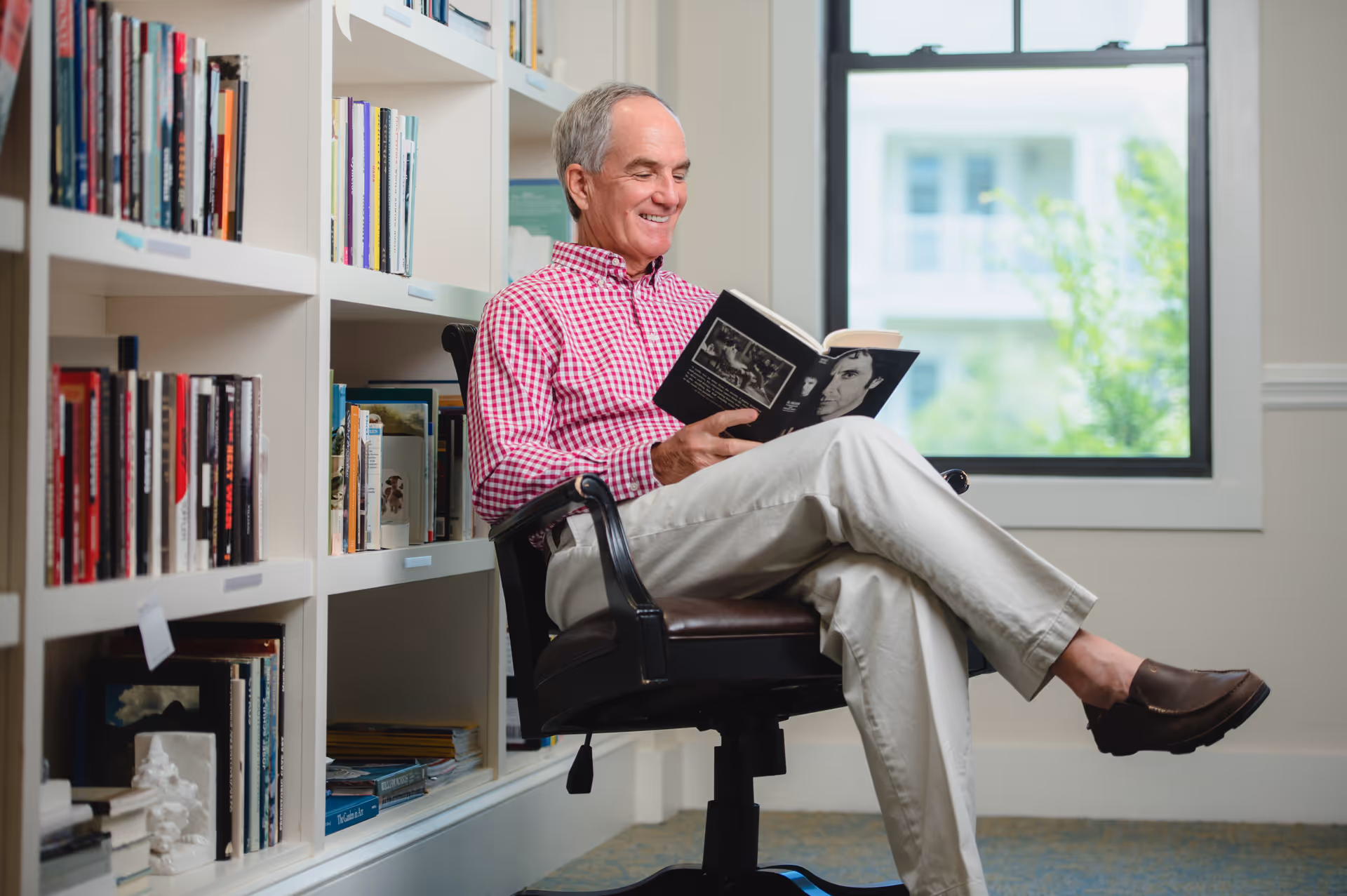 An elderly man wearing a red checkered shirt and beige pants is sitting on a black office chair in a library or reading room. He is smiling and reading a book. Behind him are white bookshelves filled with books, and there is a window with a view of greenery outside.