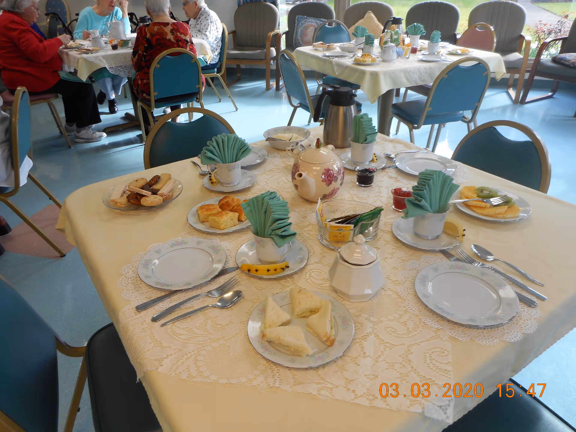 A dining room in a senior living facility with tables set for tea and snacks. The tables have lace tablecloths and are arranged with plates of sandwiches, pastries, fruit, teapots, cups with folded napkins, and utensils. Several elderly people are seated at a table in the background, engaged in conversation.