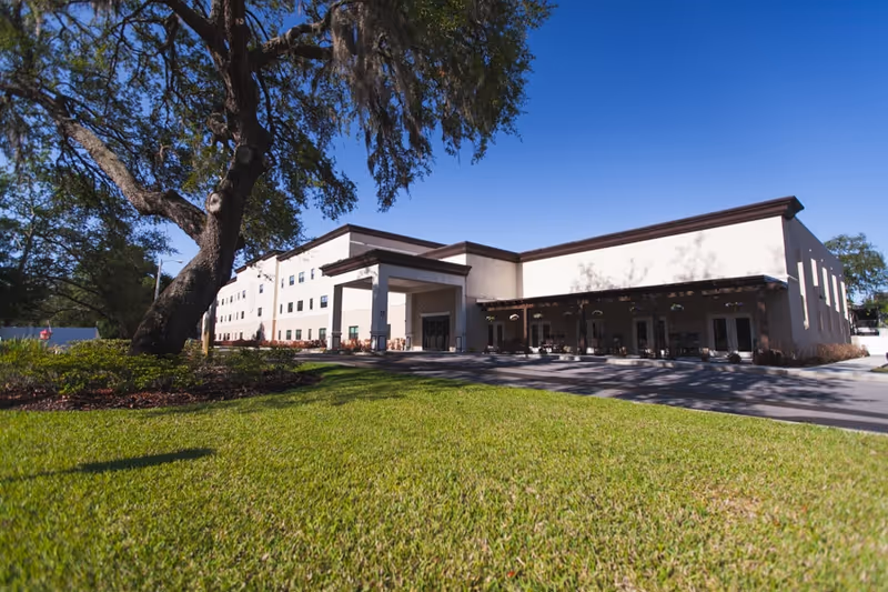 Exterior view of a large senior living facility building with a covered entrance, surrounded by green grass and trees under a clear blue sky.