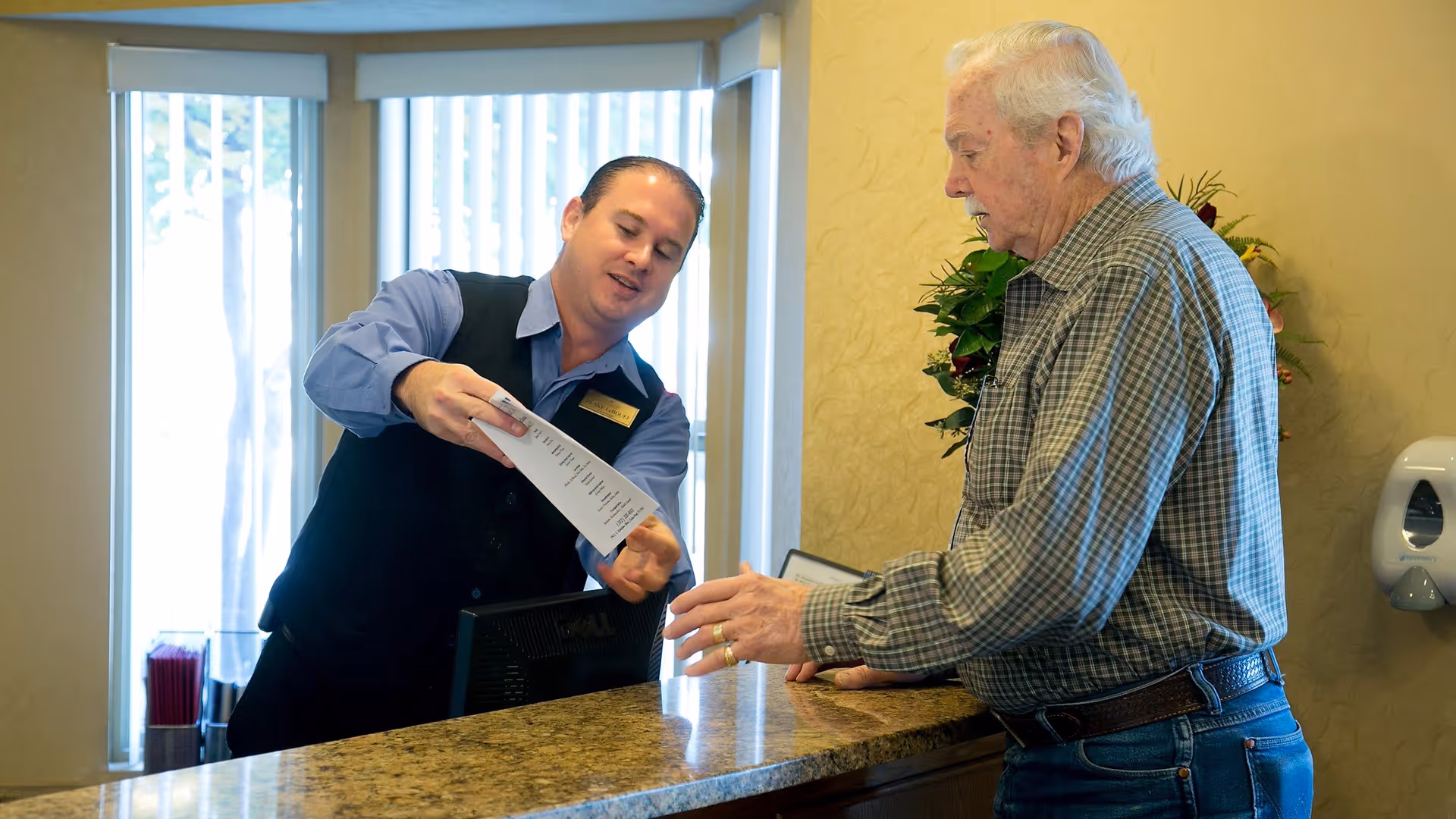 A staff member at Lakeline Oaks Retirement Community is assisting an elderly man at a reception desk, showing him a document. The setting is a well-lit interior with large windows and a granite countertop.