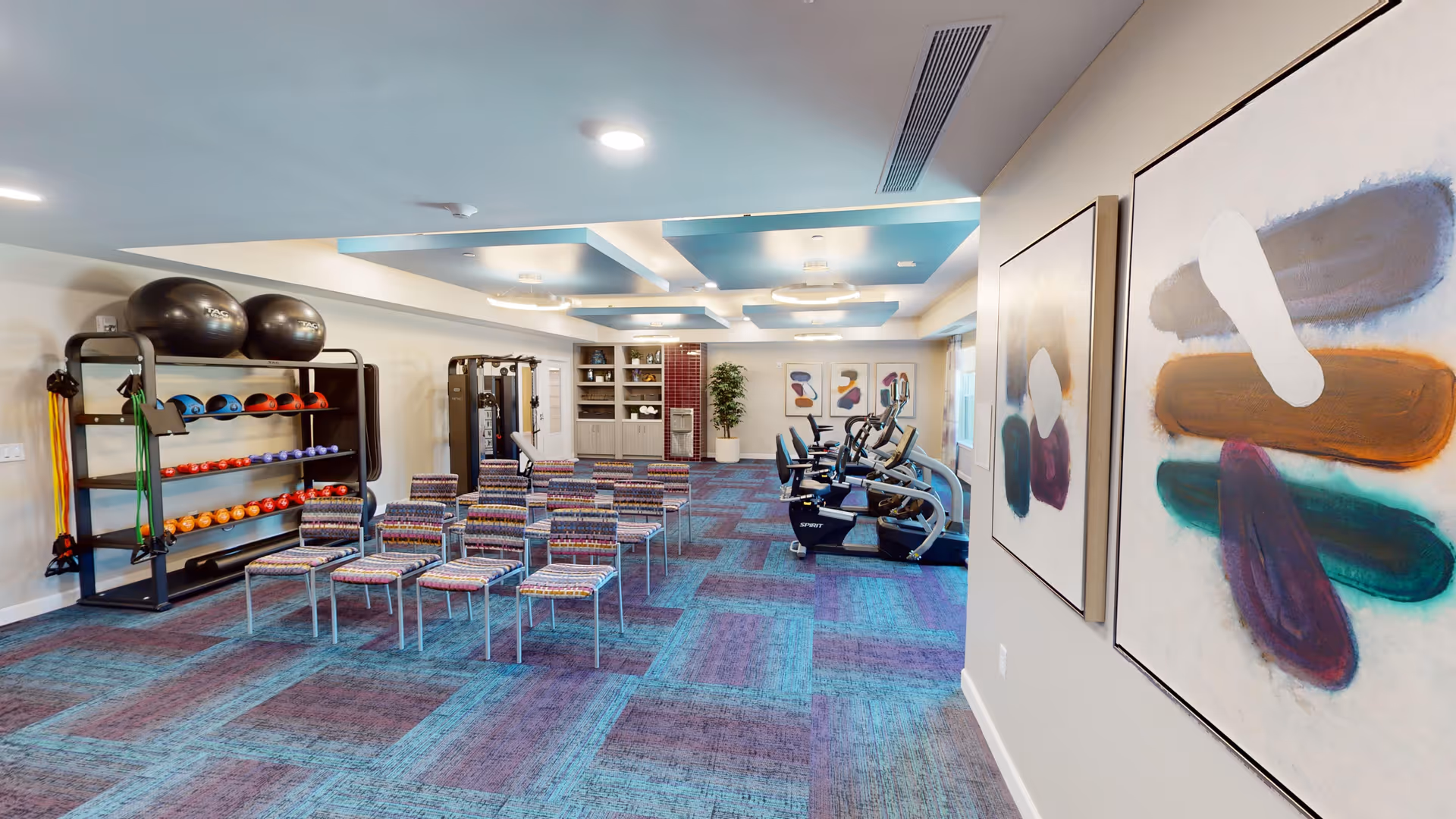 Interior fitness room with rows of chairs, exercise bikes, a rack of exercise balls and weights, and colorful wall art.