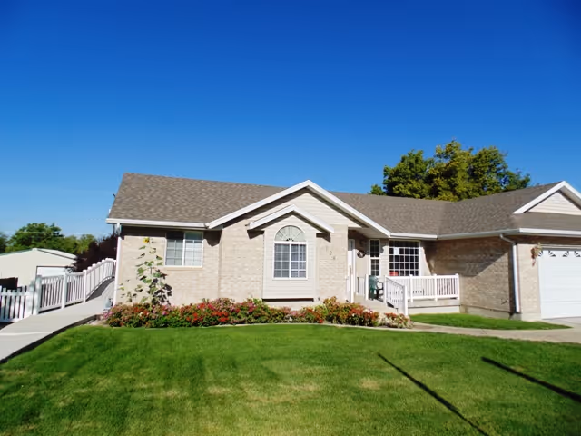 Single-story brick building with a gray shingled roof, white trim, and a small porch with white railings. There is a well-maintained green lawn and flower beds in front of the building under a clear blue sky.