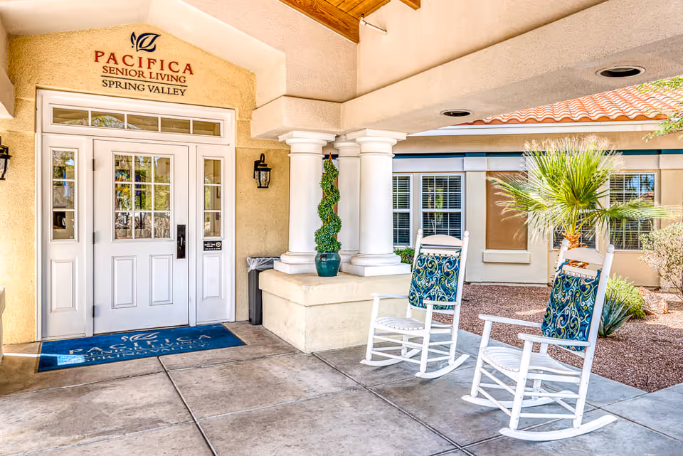Entrance to Pacifica Senior Living Spring Valley with white double doors, two white rocking chairs with blue patterned cushions on the right, a potted topiary plant, and a covered walkway with columns. The exterior walls are beige with windows and a small landscaped area with a palm tree and other plants.