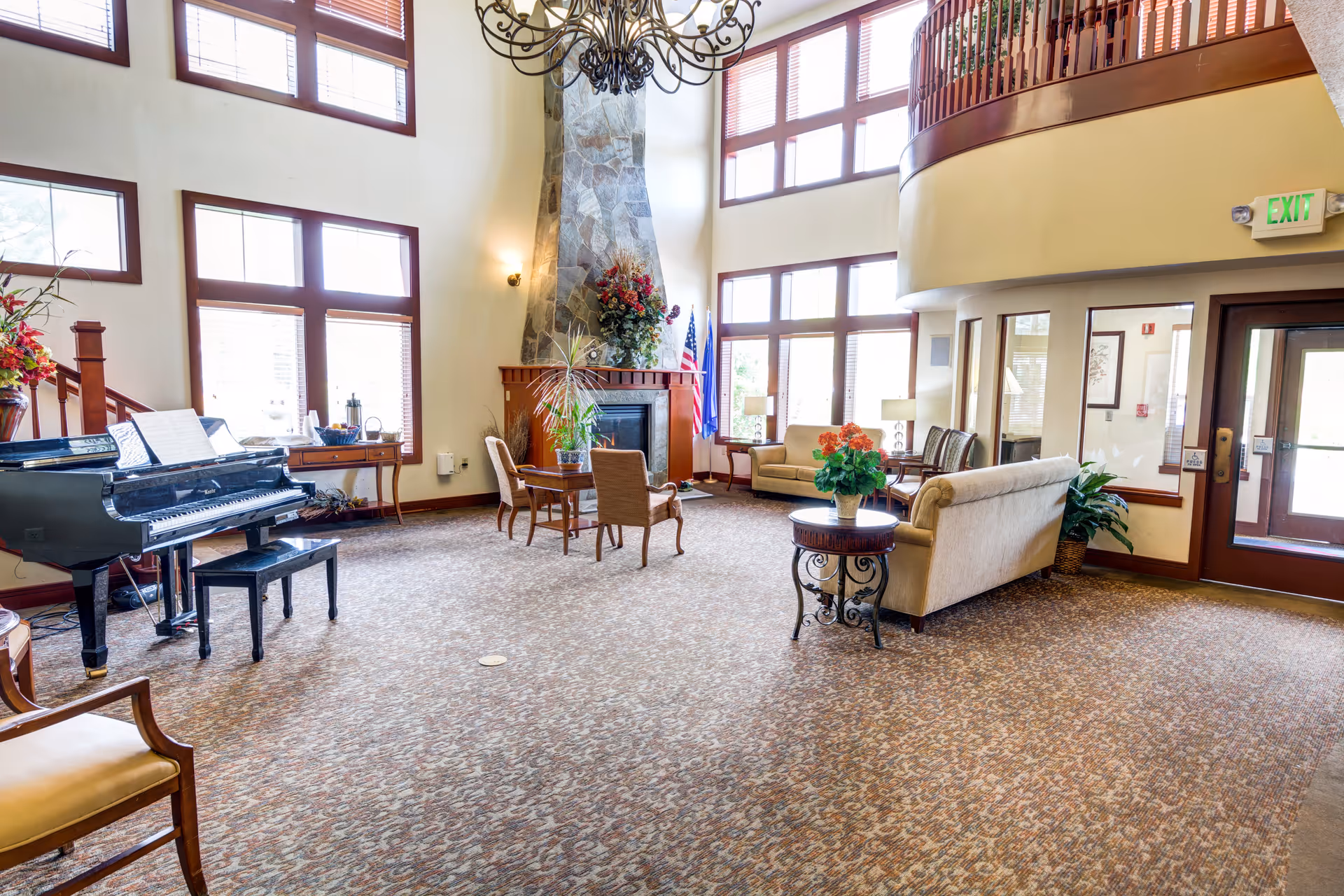 A spacious and well-lit living room area with large windows and high ceilings. The room features a stone fireplace decorated with flowers, several upholstered chairs and sofas arranged around small tables, a black grand piano with sheet music, and various plants and floral arrangements. There is a staircase with wooden railings and an exit door with glass panels.