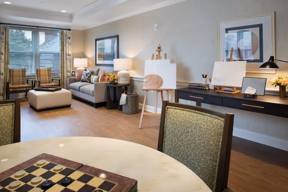 A cozy and well-lit living room area with a beige sofa, two patterned armchairs, a cushioned ottoman, and a round table with a checkers board game. The room features wooden flooring, two table lamps on side tables, framed artwork on the walls, and two easels with blank canvases and painting supplies on a long black console table.