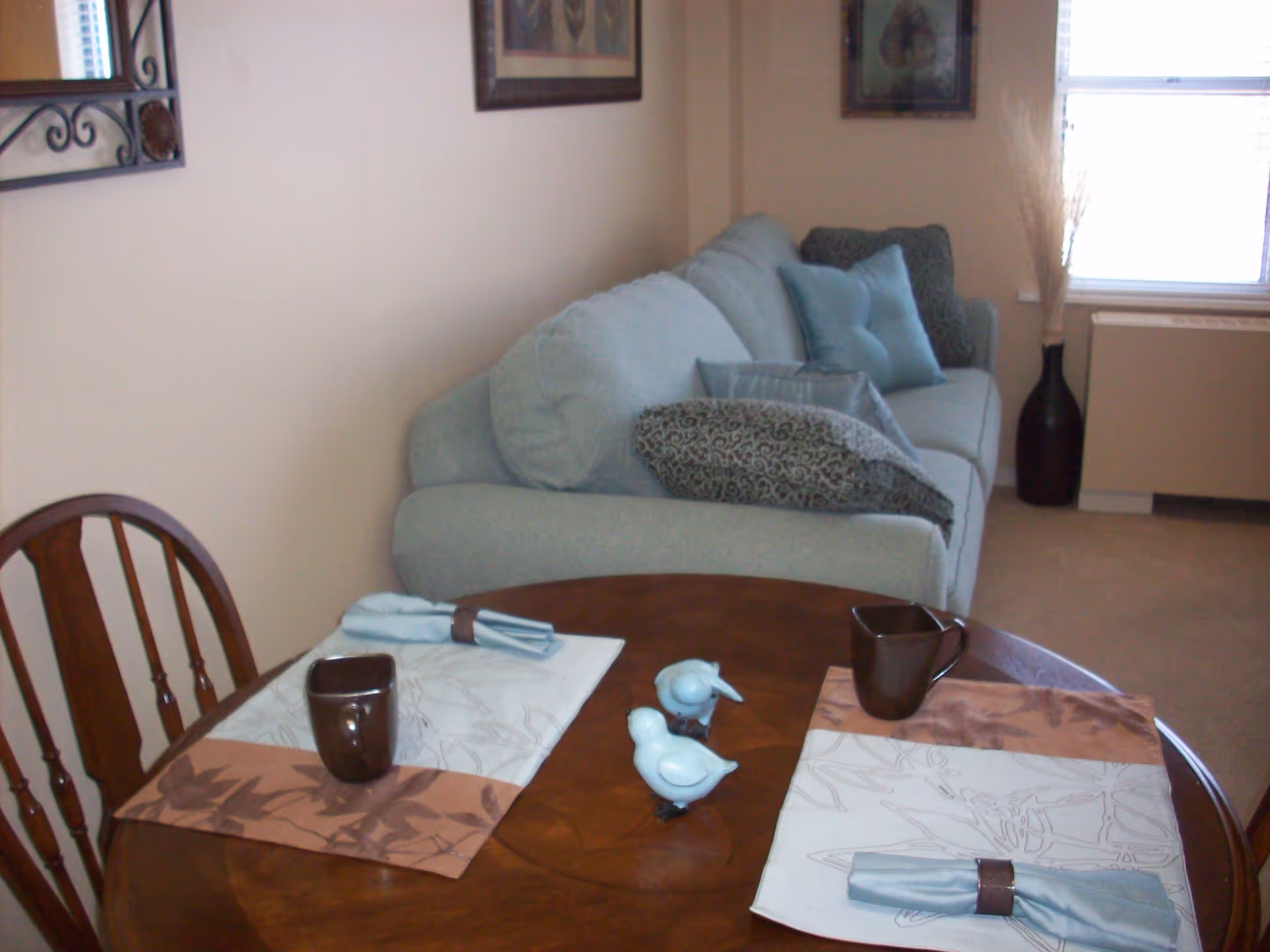 A cozy living area with a light blue sofa adorned with multiple cushions, a wooden round table set with two placemats, brown mugs, and folded napkins with napkin rings. Two small decorative bird figurines are placed in the center of the table. The room has beige walls, a window with blinds, a large dark vase with decorative sticks, and framed artwork on the walls.