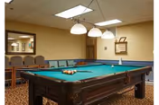 A pool table in a carpeted recreation room with hanging pendant lights and chairs along the walls.