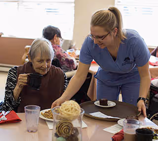 A nurse in blue scrubs smiling and serving food to an elderly woman seated at a dining table in a communal dining area. Other elderly residents are visible in the background, sitting and eating.