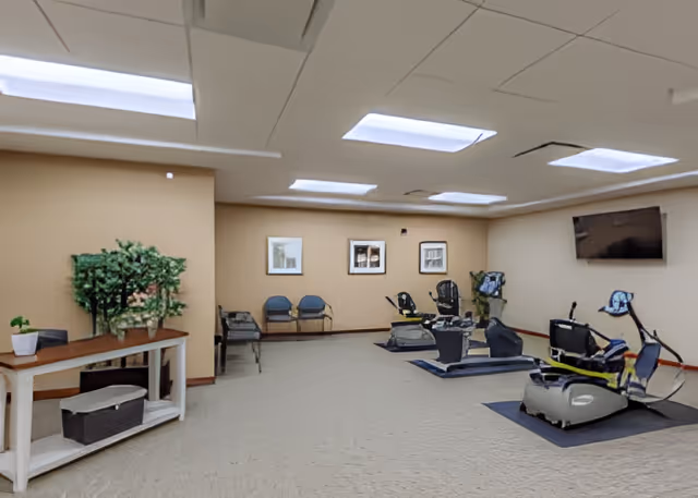 Carpeted interior activity room with several recumbent exercise bikes, seating along the back wall, a wall-mounted TV, and a table with plants.