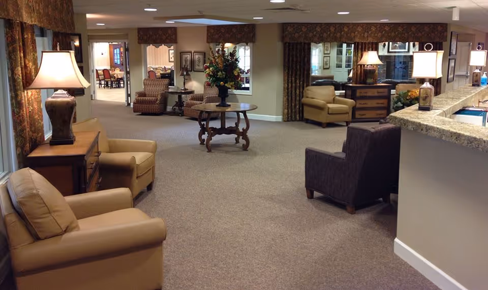 A spacious and warmly lit senior living facility common area with comfortable armchairs, side tables with lamps, a round table with a floral arrangement, and a granite countertop reception or service desk on the right side. The room has carpeted floors, patterned curtains, and framed artwork on the walls.
