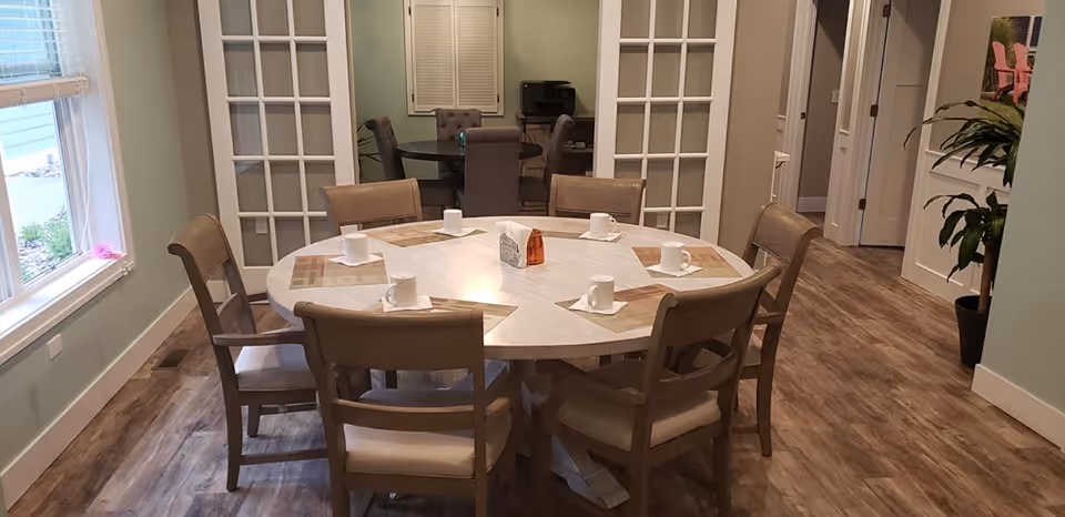 A round dining table set with six chairs in a senior living facility. Each place setting has a placemat, a white cup, and a napkin. The room has wood flooring, light-colored walls, and a window on the left side. In the background, there is another dining area with a rectangular table and chairs, separated by glass-paneled double doors. A potted plant is visible on the right side near a hallway.