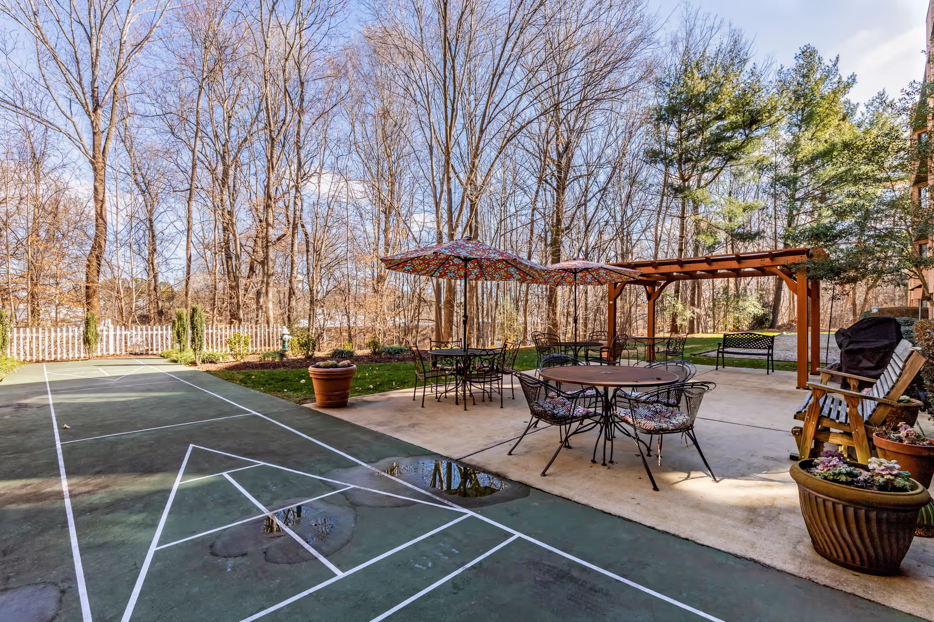 Outdoor patio area with round tables and chairs under colorful umbrellas, a wooden pergola, potted plants, and a shuffleboard court. The setting is surrounded by leafless trees and a white picket fence under a partly cloudy sky.