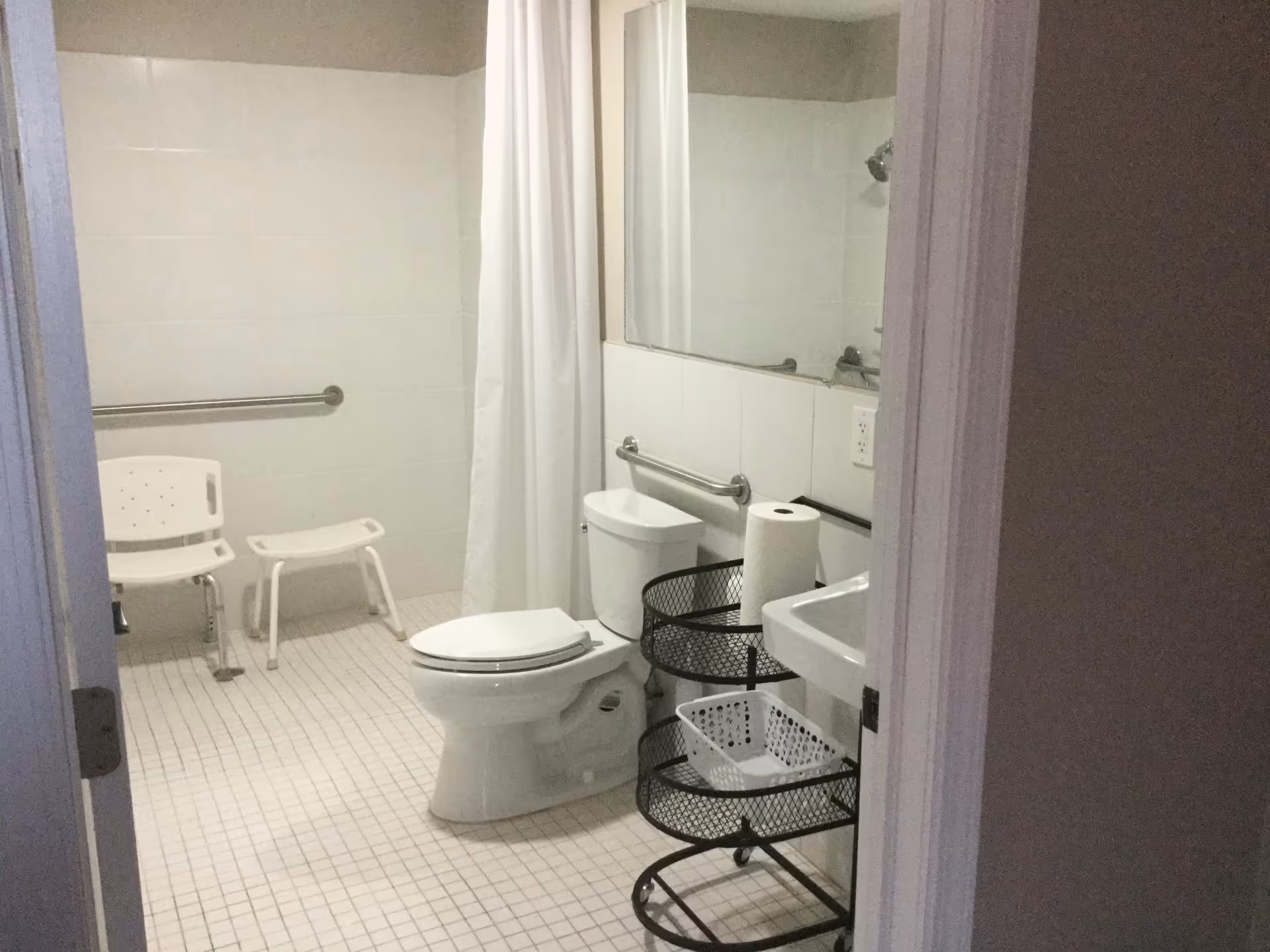 A bathroom with white tiled floor and walls, featuring a toilet, a white shower chair, a small white stool, a shower area with a white curtain, a sink, a large mirror, and a black metal rolling cart holding a roll of paper towels and a white basket.