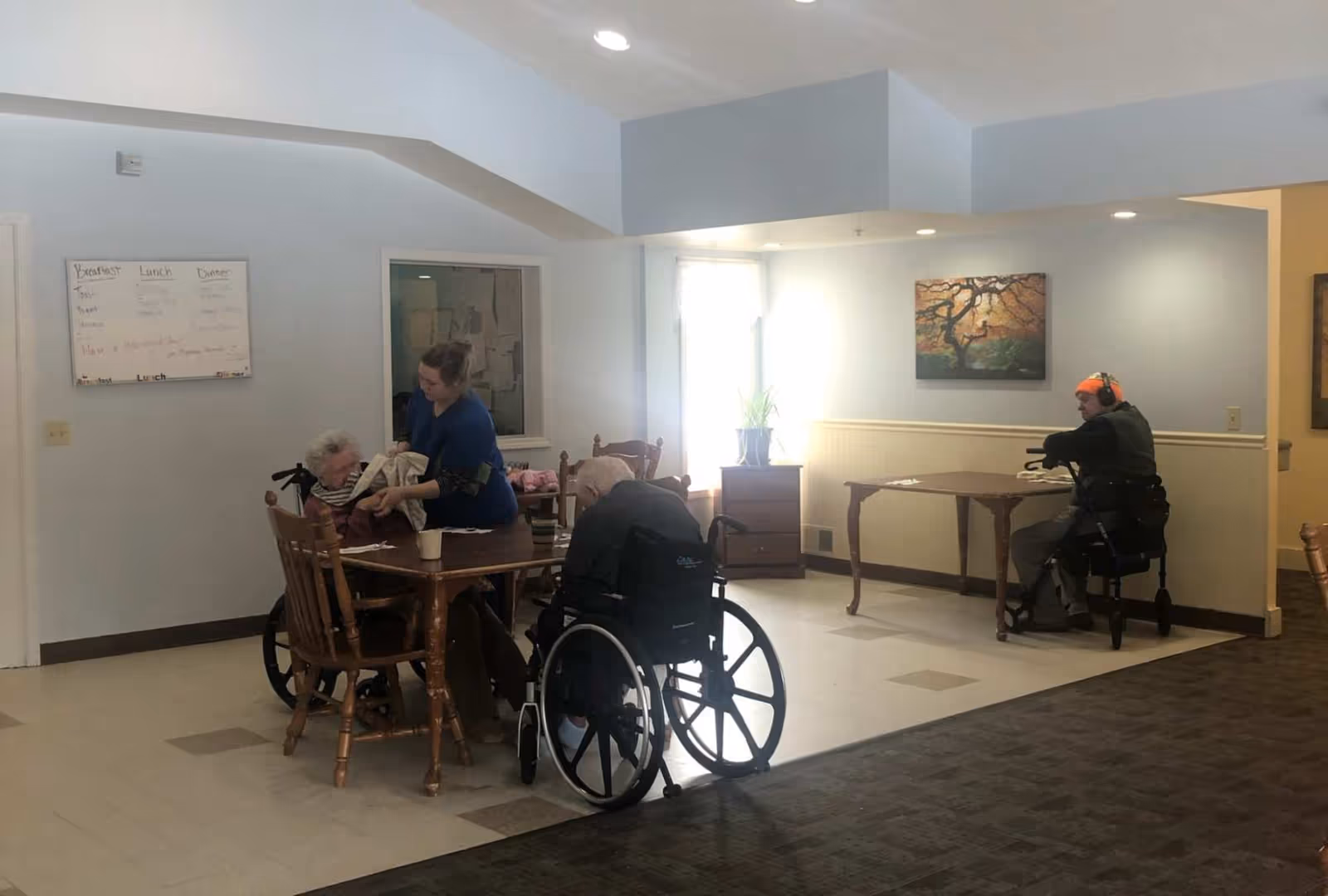 Caregivers and residents seated at tables in a communal dining/activity area of a senior living facility.