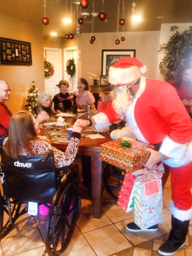 A festive holiday gathering in a senior living facility dining room with Christmas decorations hanging from the ceiling. A person dressed as Santa Claus is handing a gift to a woman in a wheelchair, while other seniors and staff members sit around a table smiling and enjoying the celebration.
