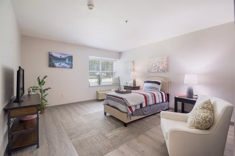 Bright furnished bedroom featuring a single bed with striped bedding, an armchair, TV on a stand, nightstands and a window.
