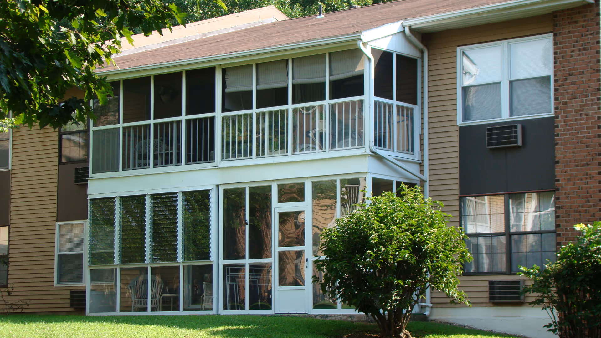 Exterior view of a residential building with a two-story screened-in porch. The building has beige siding and brick sections, with several windows and air conditioning units. There is green grass and bushes in the foreground.
