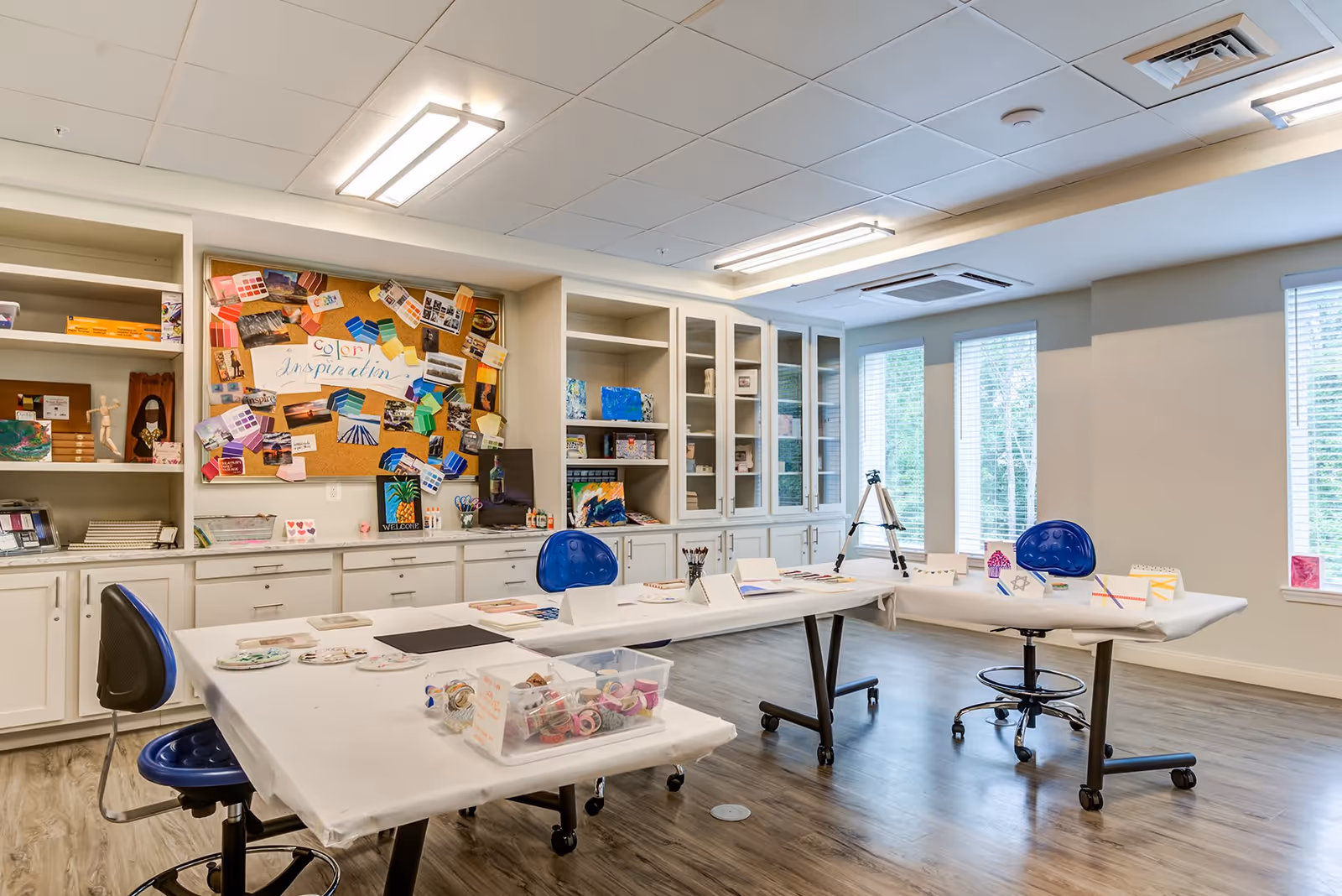 A bright and spacious art and craft room with two tables covered in white paper, blue swivel chairs, and various art supplies including paintbrushes, paints, and decorative tapes. The back wall features built-in white cabinets and shelves filled with art materials and a colorful bulletin board labeled 'Color Inspiration' displaying paint swatches and photos. Large windows on the right side let in natural light.