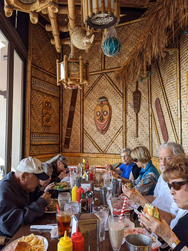 A group of elderly people sitting around a long wooden table in a tiki-themed restaurant, eating various meals including burgers and fries. The walls are decorated with bamboo, woven panels, and tribal masks. Condiments and drinks are on the table.