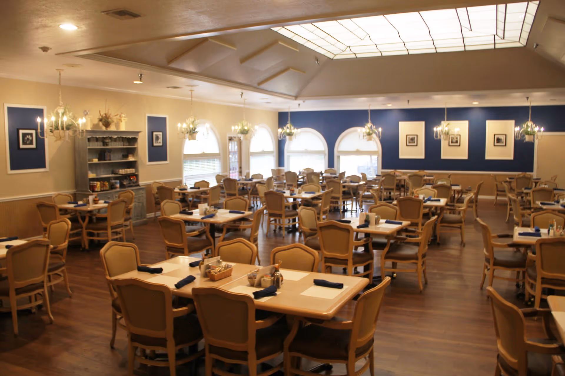 A spacious dining room with multiple tables and chairs arranged neatly. Each table has place settings with napkins and condiments. The room features wooden flooring, chandeliers hanging from the ceiling, and large arched windows letting in natural light. The walls are decorated with framed pictures and blue accent panels.