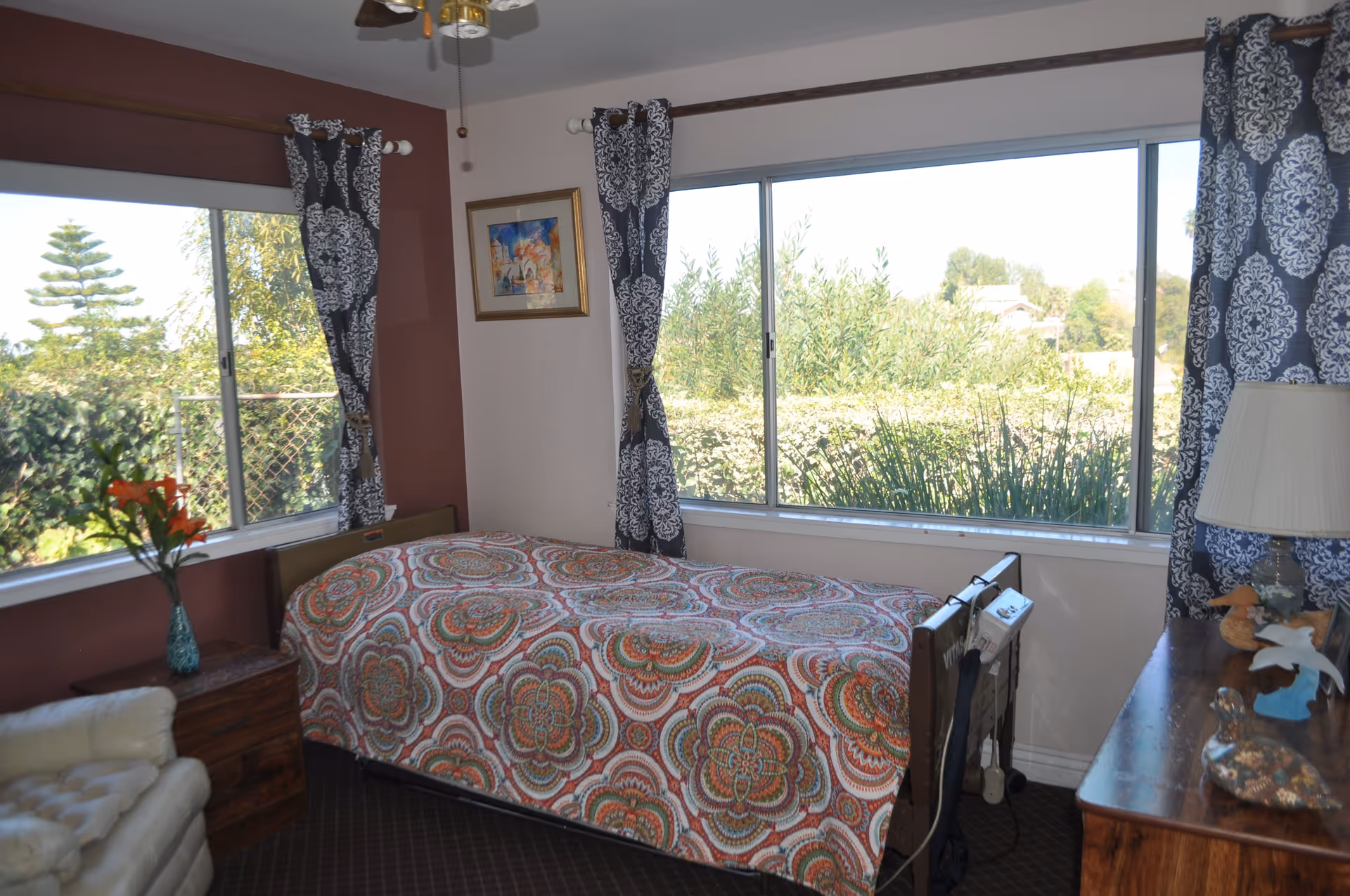 A bedroom with a single bed covered in a colorful patterned bedspread. The room has large windows with patterned curtains, letting in natural light and showing greenery outside. There is a wooden dresser with a lamp and decorative items, a small wooden nightstand with a vase of flowers, and a white armchair in the corner.