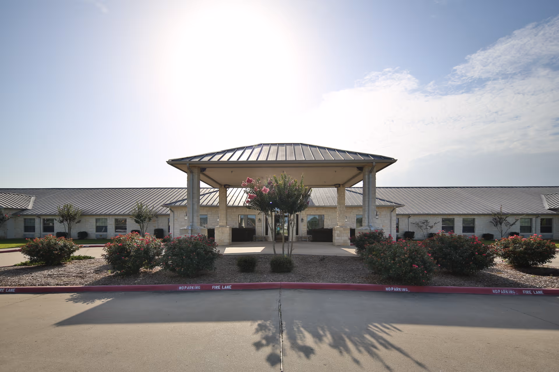 Front entrance of a single-story nursing and rehabilitation center with a covered porte-cochere, landscaped beds, and a metal roof under a bright sky.