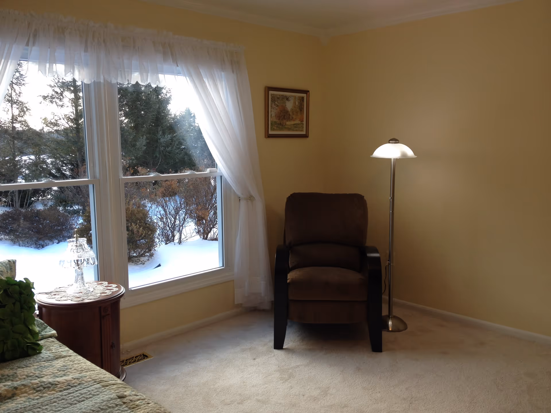 Sunlit bedroom sitting area with a brown recliner, floor lamp, small round bedside table and a large window showing snow outside.