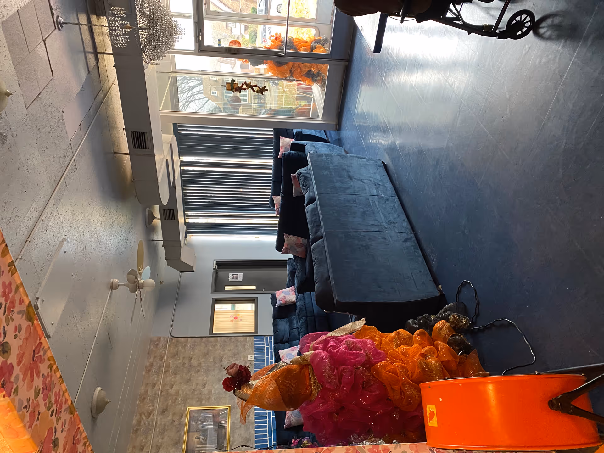 Interior view of a living room area in a senior living facility with large windows letting in natural light. The room features dark blue sofas with pink pillows, a ceiling fan, and decorative orange and pink mesh decorations near the entrance. A wheelchair is partially visible on the right side of the image.