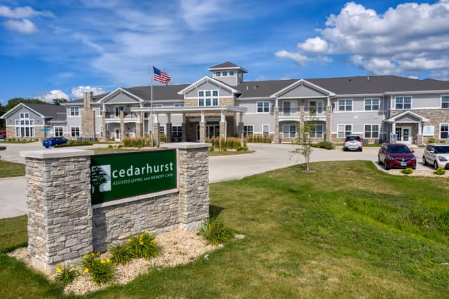 Exterior view of Cedarhurst assisted living and memory care facility in Yorkville, showing a large two-story building with stone and siding facade, an American flag on a flagpole, a driveway with parked cars, and a stone sign with the Cedarhurst logo in the foreground under a partly cloudy blue sky.