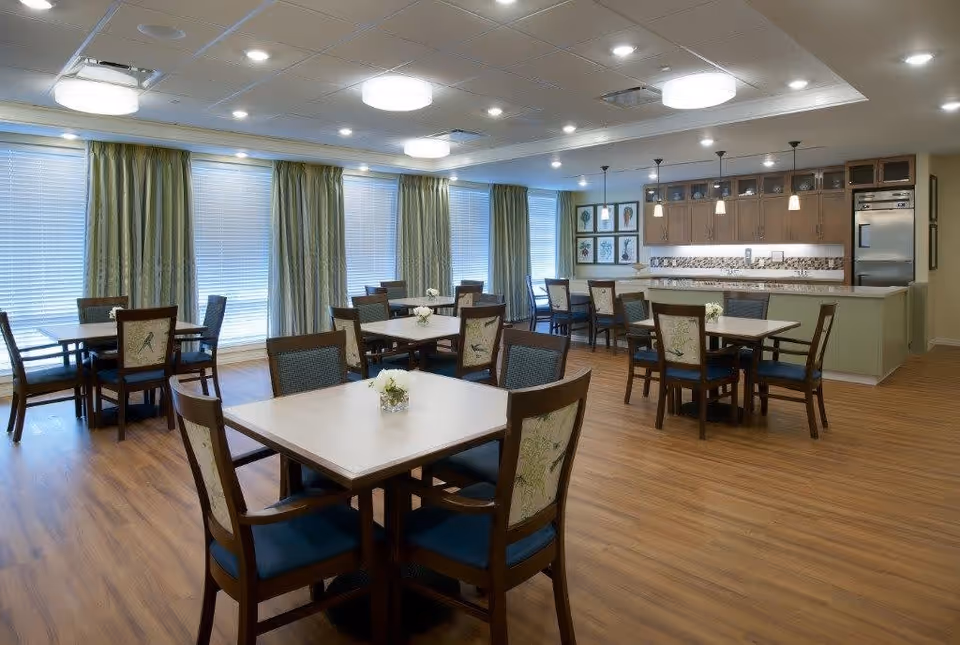 A spacious dining room with multiple square tables each surrounded by four wooden chairs with patterned backs and blue cushions. The room has large windows covered with blinds and green curtains, wooden flooring, and a kitchen area with cabinets, a countertop, and pendant lights in the background.