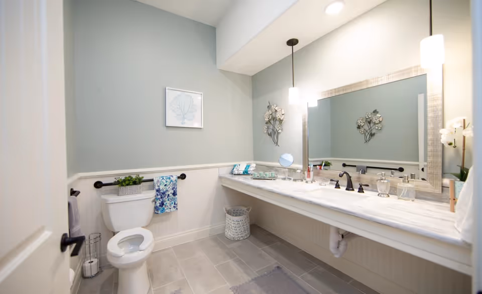 A bright accessible bathroom with a toilet, long marble countertop vanity and large mirror, pendant lights and grab bars.