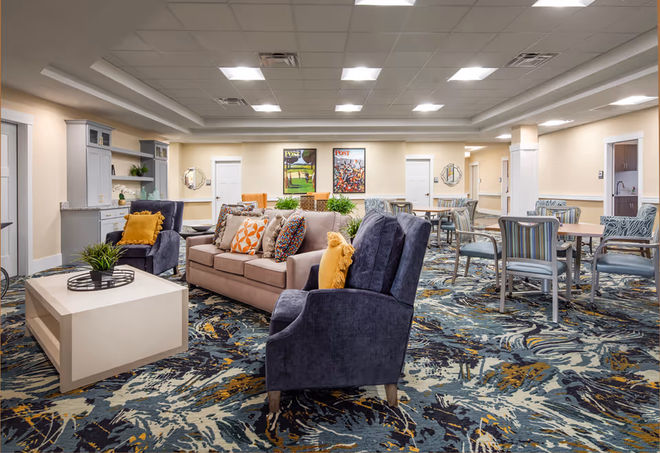 A spacious and well-lit common area in a senior living facility featuring a beige sofa with colorful cushions, two dark blue armchairs with yellow pillows, a white coffee table with a plant, and several tables with chairs in the background. The room has a patterned carpet, light-colored walls, ceiling lights, and framed artwork on the far wall.