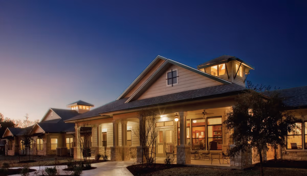 Exterior view of a single-story assisted living facility building at dusk with warm interior lights glowing through the windows. The building features a covered porch with rocking chairs and a peaked roof with a cupola on top.