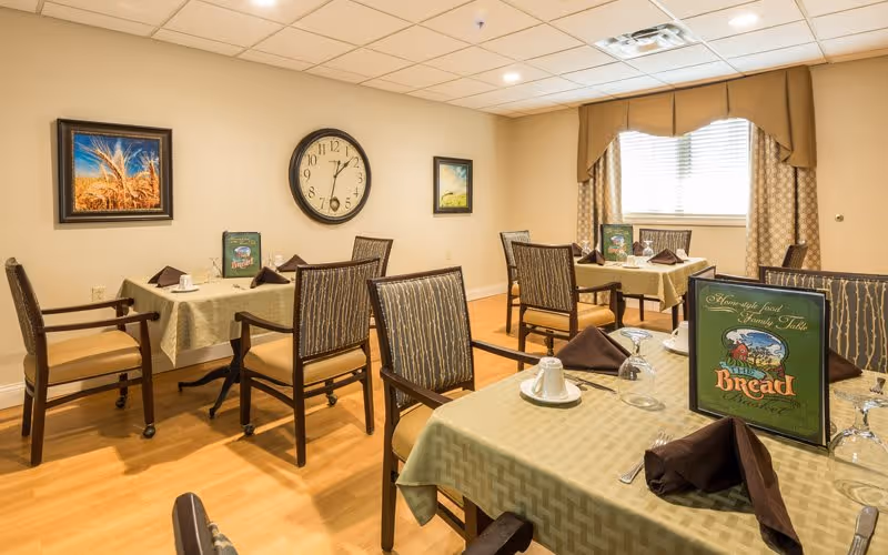 A dining room in a senior living facility with several tables covered in green tablecloths, each set with brown napkins, cups, and glasses. The room has wooden chairs with patterned upholstery, a large clock on the wall, framed artwork, and a window with beige curtains allowing natural light.