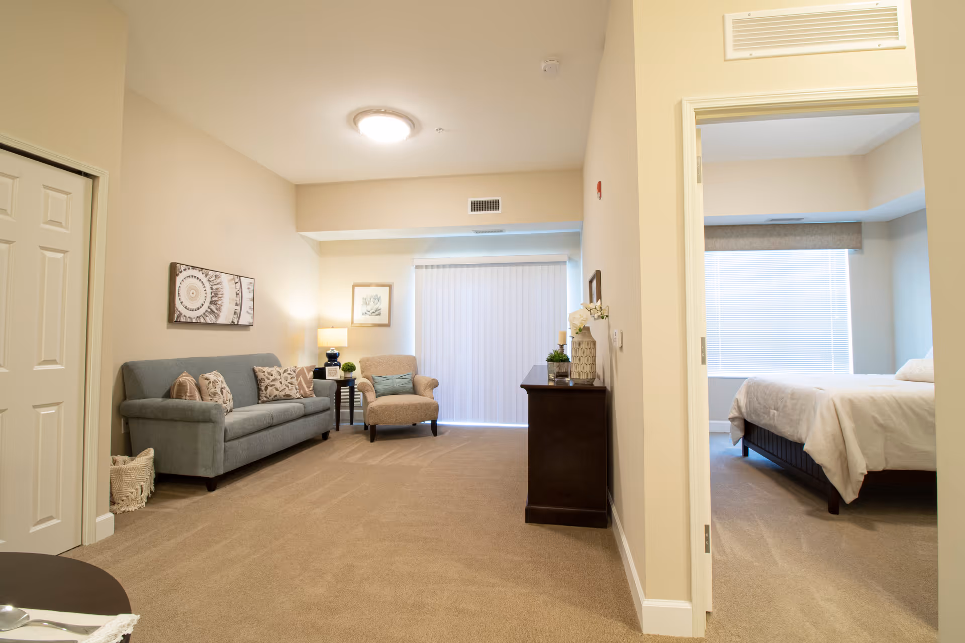 A bright and clean living area with a gray sofa adorned with patterned pillows, a beige armchair, a side table with a lamp, and a dark wooden cabinet with decorative items. To the right, an open doorway reveals a bedroom with a neatly made bed and a large window with blinds.