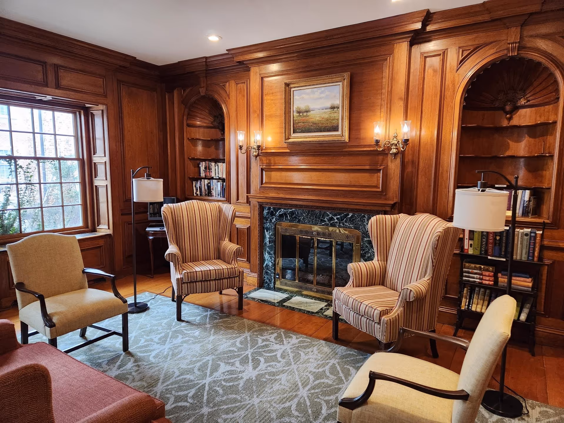 A cozy living room with wood-paneled walls and a fireplace with a marble surround. There are four upholstered chairs arranged around a patterned area rug, two floor lamps, built-in bookshelves with books, and a framed landscape painting above the fireplace. A large window lets in natural light.