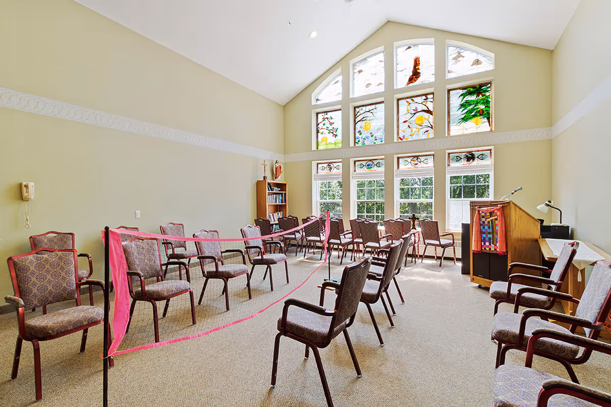 A spacious room with high ceilings and large stained glass windows letting in natural light. The room is arranged with multiple rows of padded chairs separated by a pink ribbon, facing a wooden podium with a colorful cloth draped over it. There is a bookshelf in the corner and a telephone mounted on the wall.