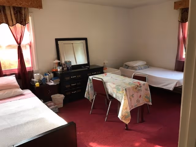 A simple bedroom with two single beds, each with pillows and bedding. Between the beds is a small table covered with a floral tablecloth and two chairs. Against the wall is a dark dresser with a mirror resting on top, along with various personal items. The room has red carpet flooring and two windows with red curtains letting in natural light.
