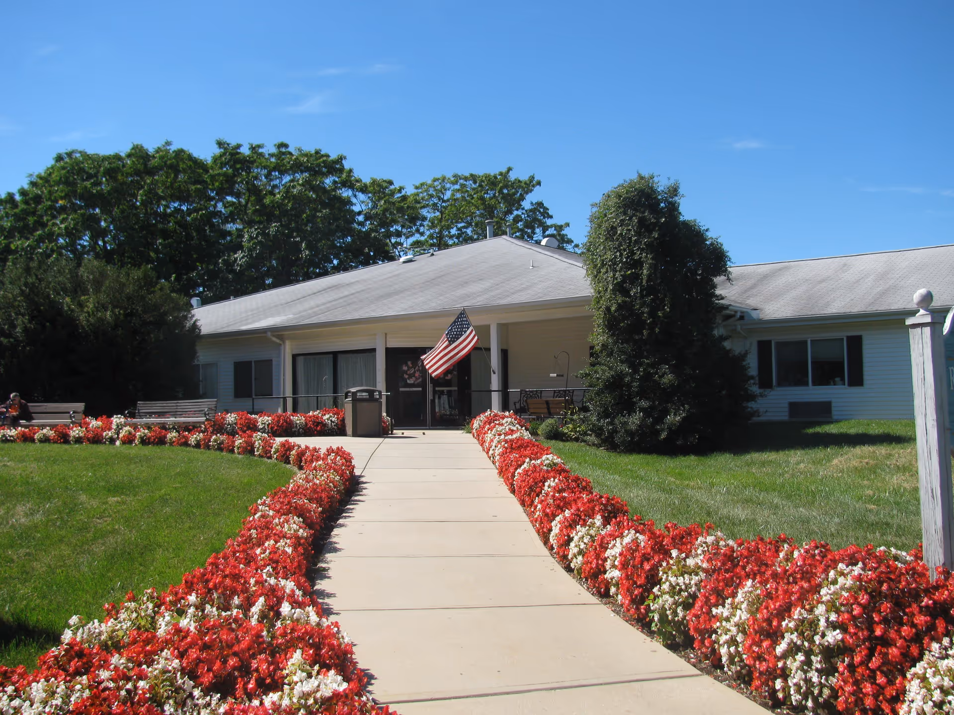 A paved walkway lined with red and white flowers leads to the entrance of a single-story building with a gray roof and white siding. An American flag is displayed near the entrance, and there are benches and greenery around the building under a clear blue sky.
