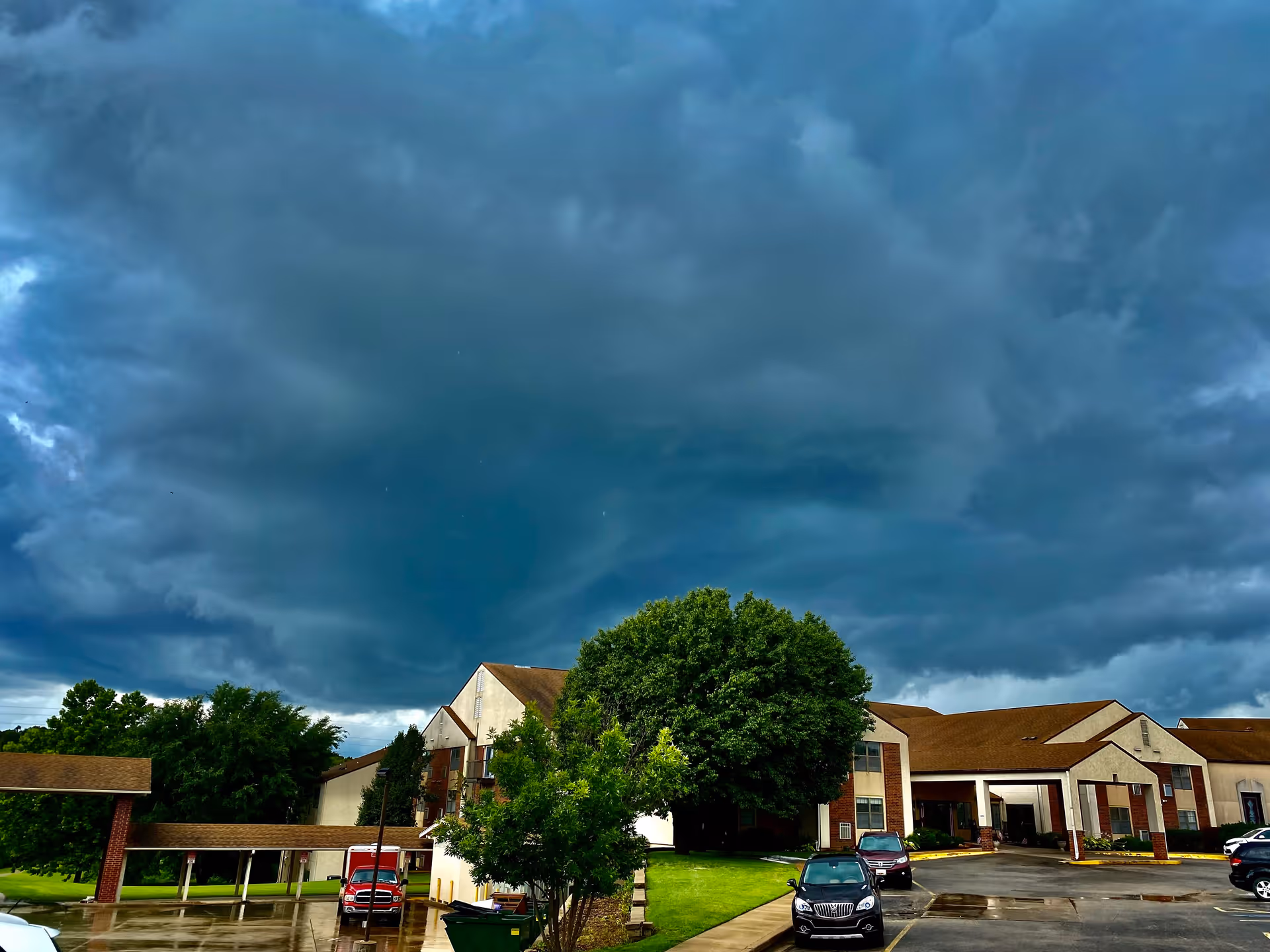 Exterior view of Green Country Village senior living facility under a dramatic cloudy sky, showing a multi-story building with brown roofs, a covered entrance, parked cars, and green trees and lawn.