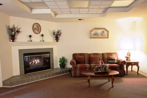 Cozy lounge area with a lit fireplace, patterned sofa, coffee table with flowers, and a side table lamp.