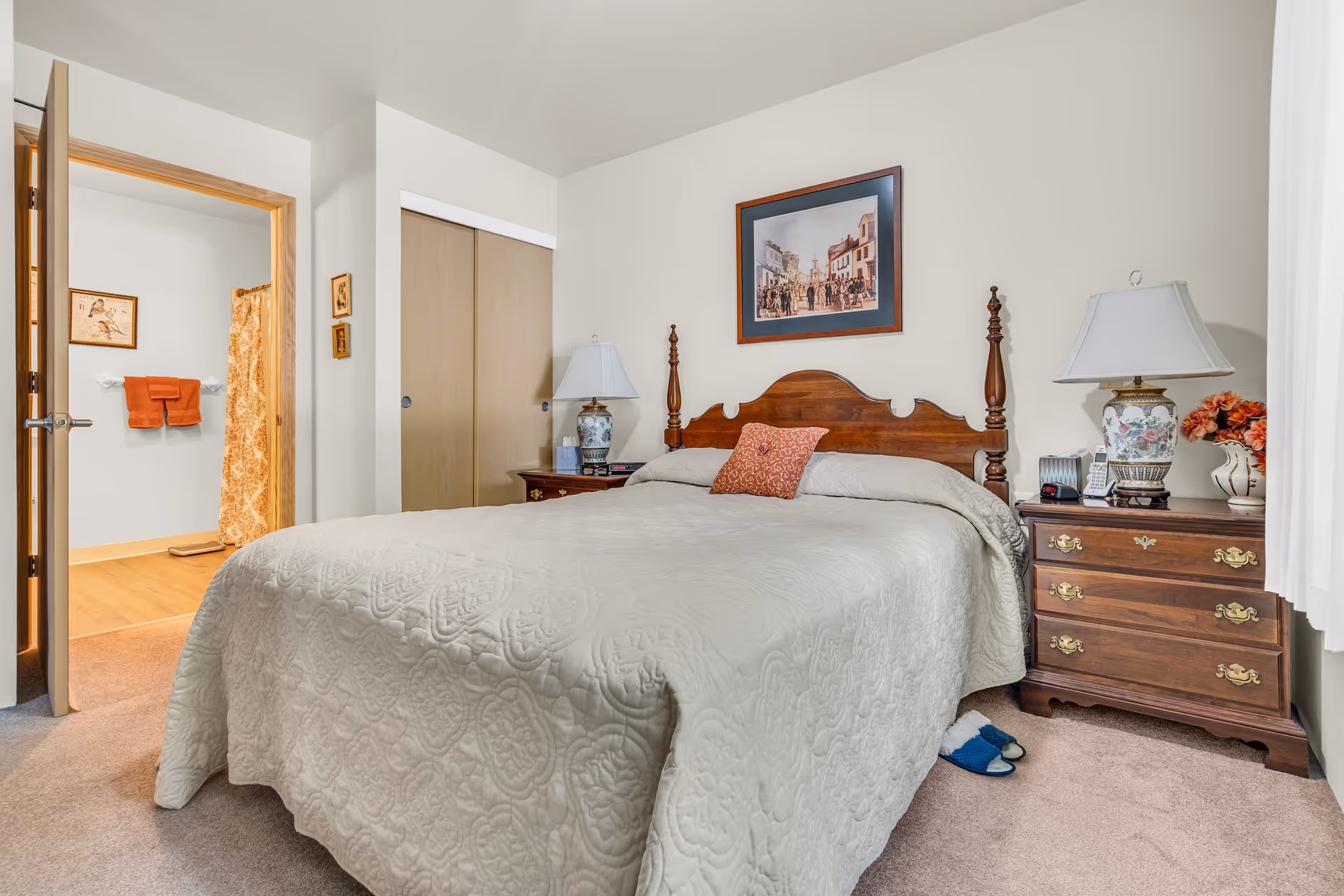 A cozy bedroom with a large bed covered in a light beige quilt. The bed has a wooden headboard and a single decorative pillow. On either side of the bed are wooden nightstands with matching lamps. The room has beige carpet and light-colored walls, with a framed picture hanging above the bed. An open door reveals a bathroom with orange towels and a shower curtain.