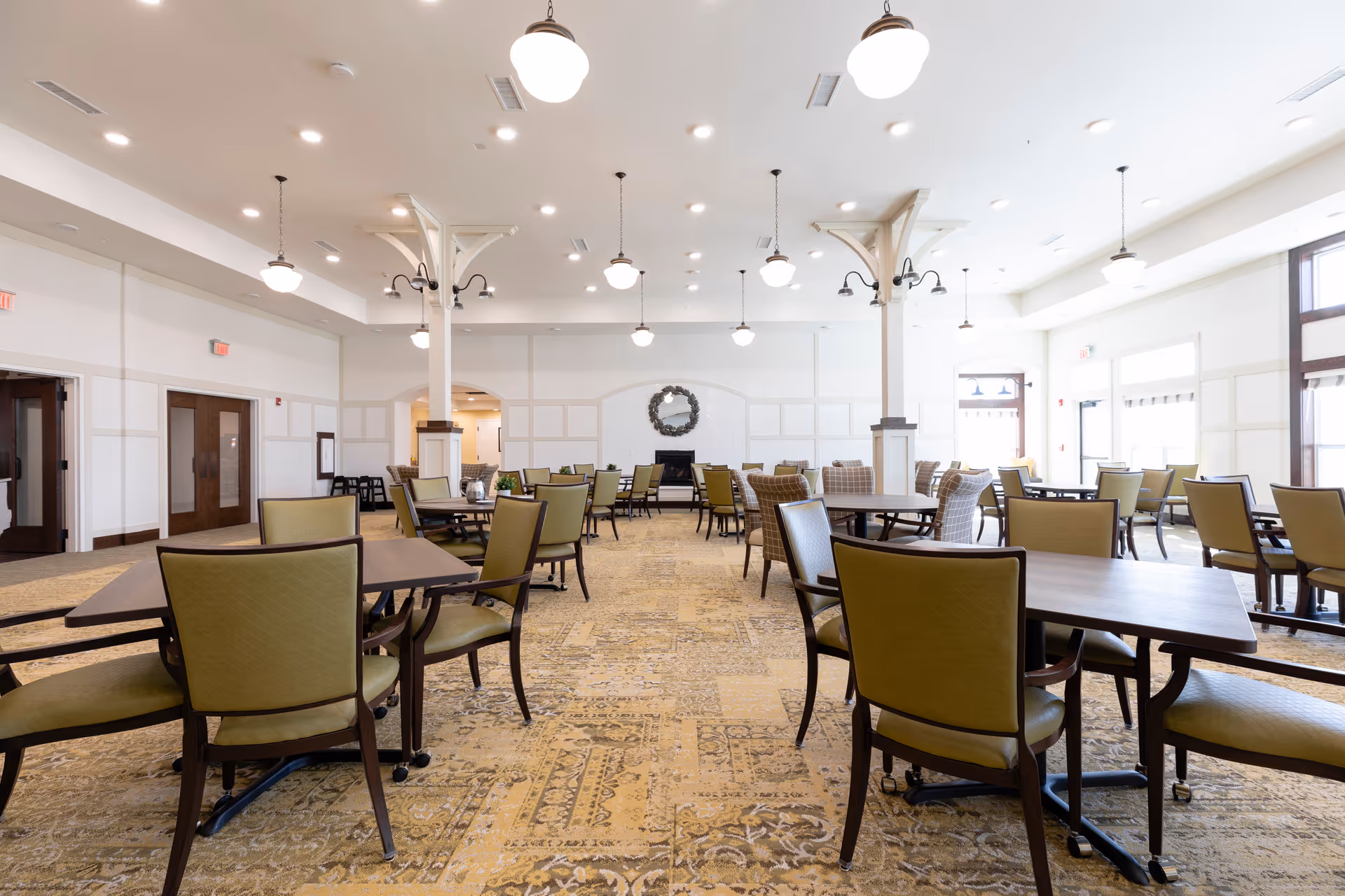 A spacious and well-lit dining room with multiple tables and green cushioned chairs arranged neatly. The room features a patterned carpet, white walls with paneling, several hanging light fixtures, and large windows allowing natural light to fill the space.