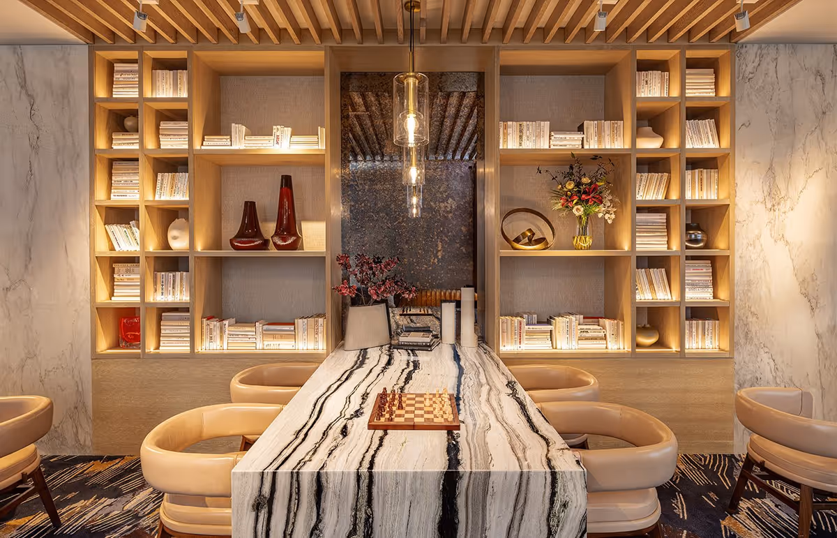 A modern, elegant interior room featuring a long marble table with black and white streaks, surrounded by beige leather chairs. The table has a chessboard set up on it. Behind the table is a large built-in wooden bookshelf filled with white-covered books and decorative items such as vases and flowers. The room has warm lighting with pendant lights hanging above the table and marble walls on the sides.