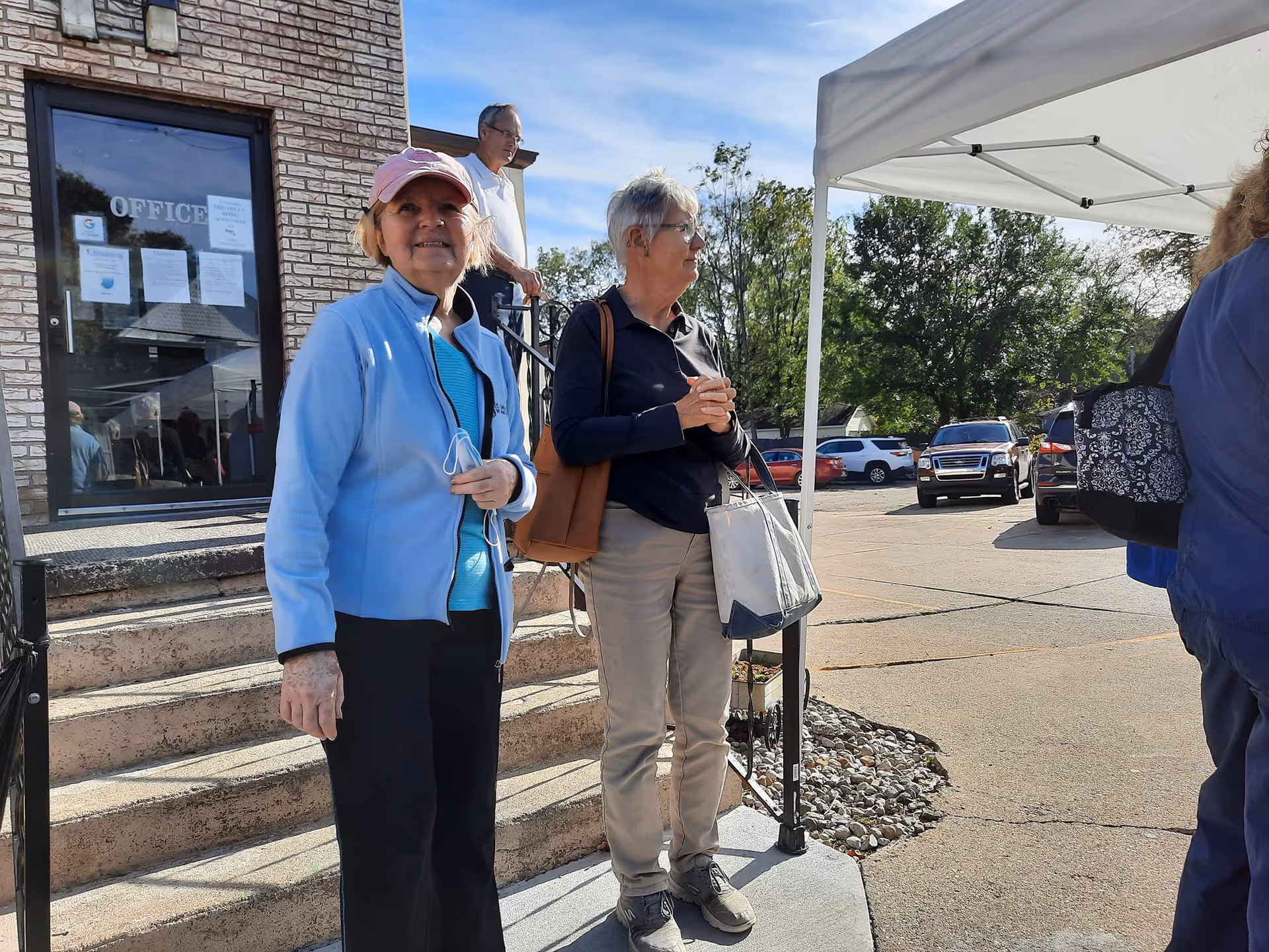 Two elderly women standing outside near the entrance of a building with a sign that reads 'OFFICE' on the door. One woman is wearing a light blue jacket and pink cap, smiling at the camera, while the other woman is wearing a dark jacket and holding a white bag, looking to the side. A man is seen descending stairs in the background, and several parked cars and trees are visible under a clear sky.