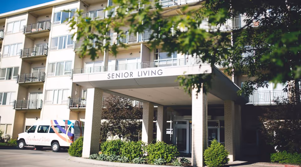 Entrance of a multi-story senior living building with a covered drop-off reading 'SENIOR LIVING' and a shuttle van parked nearby.