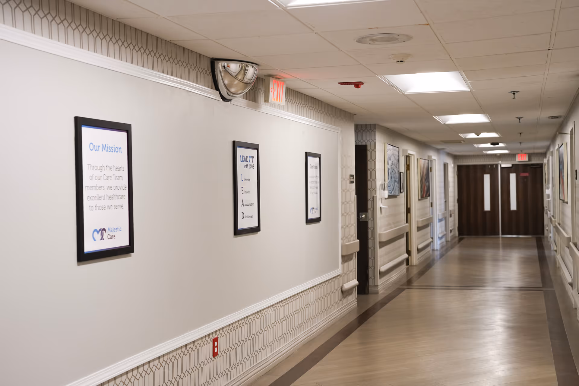 Well-lit interior hallway in a senior living facility with framed posters along the wall and double doors at the far end.