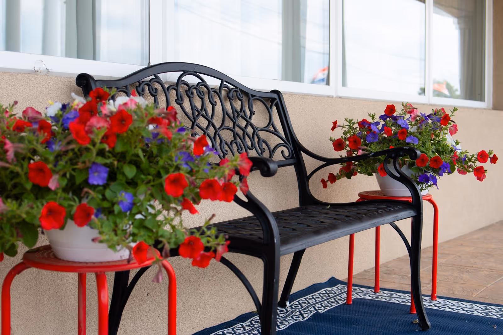 Black decorative metal bench on a porch with potted red and purple flowers on red stools beneath windows.