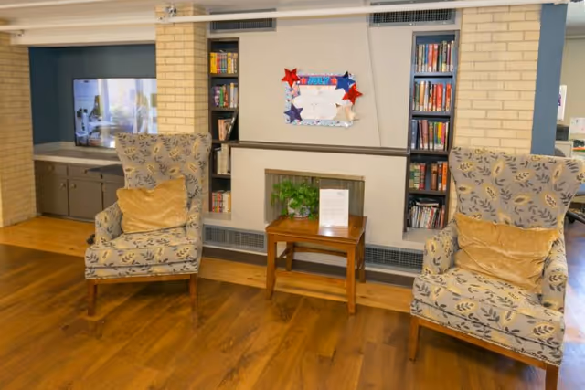 Two patterned upholstered armchairs with gold pillows flank a small wooden table in a cozy seating area with built-in bookshelves, a bulletin board, and a TV.